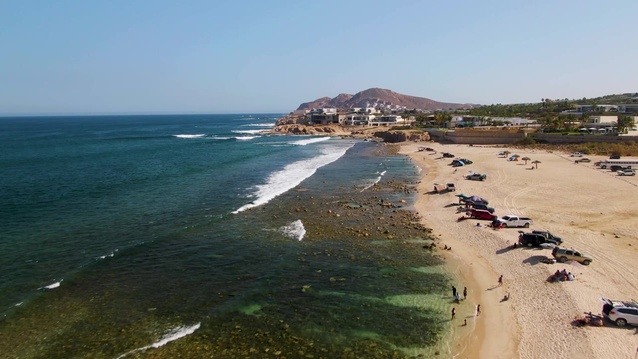 Aerial coastline frame from El Tule Beach in Los Cabos showing a stronger white surf band crossing the rocky shallows, scattered vehicles on the sand, small visible beachgoers, and hills behind coastal buildings.