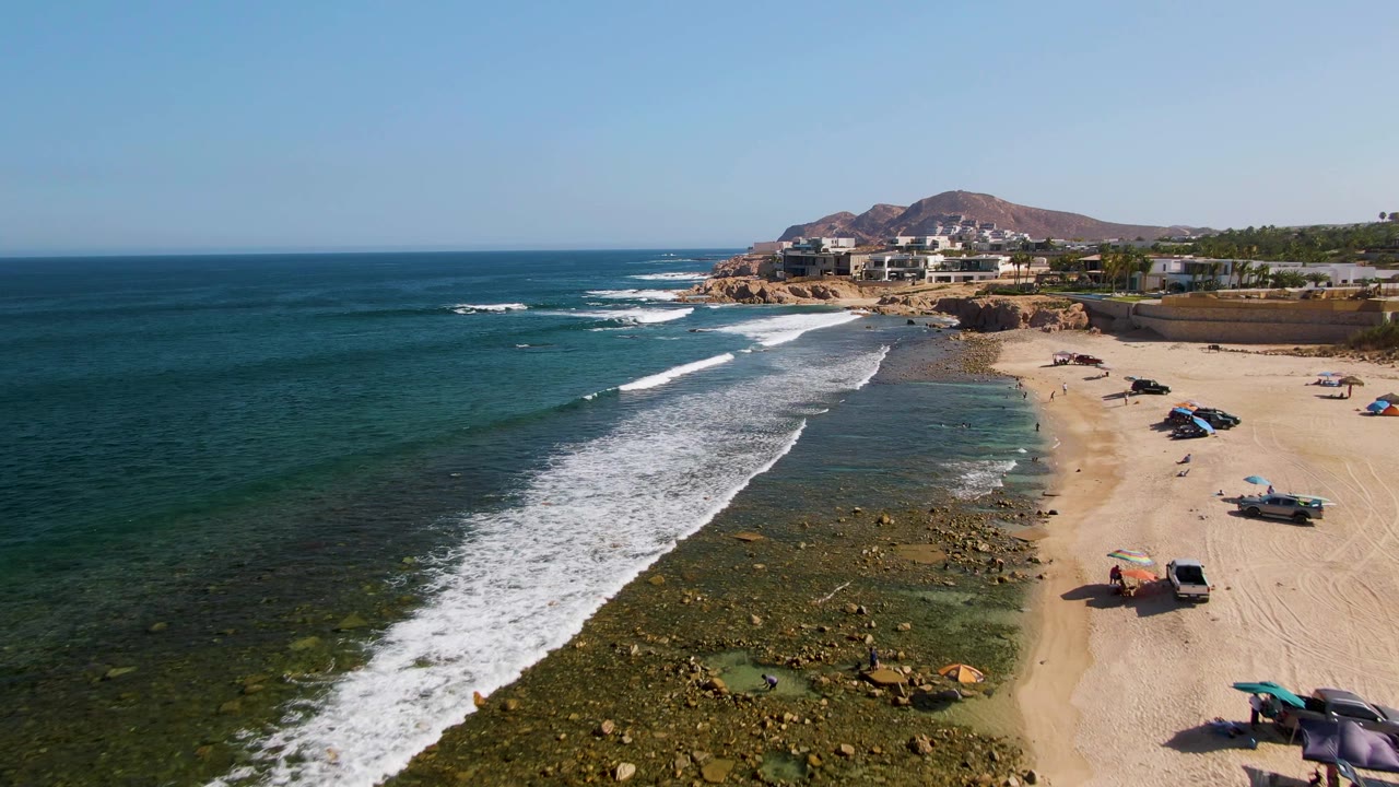 Aerial view over El Tule Beach in Los Cabos, with white waves entering from the left, rocky shallows near shore, parked vehicles on the sand, and low buildings beyond the beach.