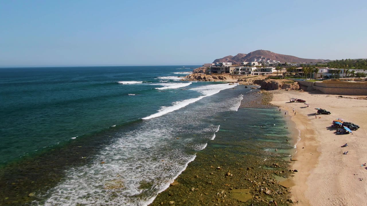 Wide aerial view at El Tule Beach in Los Cabos, showing white waves over shallow rocky water, a broad sand strip, scattered parked vehicles, small beach structures, and hillside buildings.