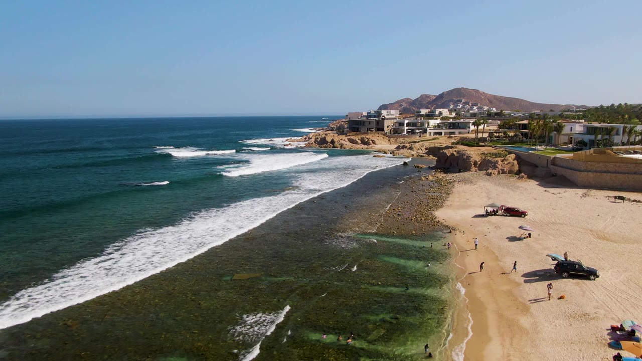 Aerial view of El Tule Beach in Los Cabos with white waves crossing rocky shallows, a sandy beach with vehicles, scattered people near the water, and beachfront buildings on the bluff.