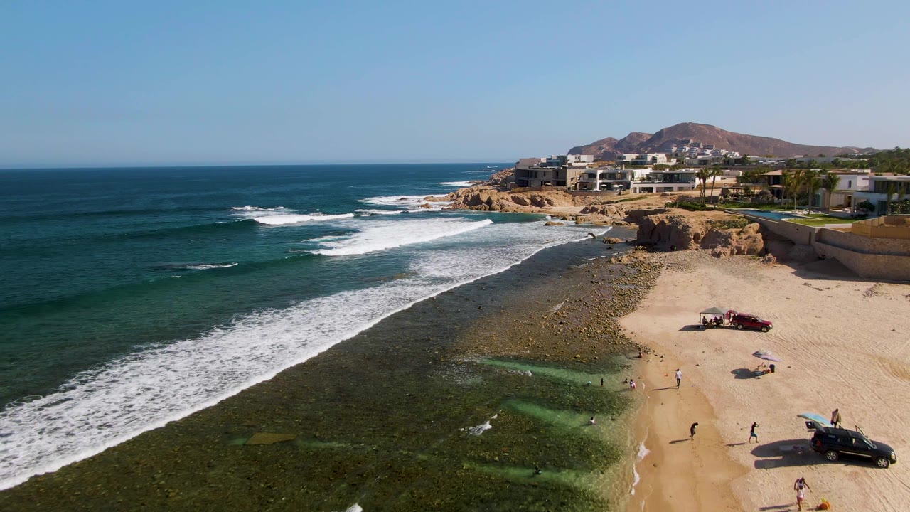 Drone view of surf rolling toward a sandy beach with two vehicles and beachgoers.