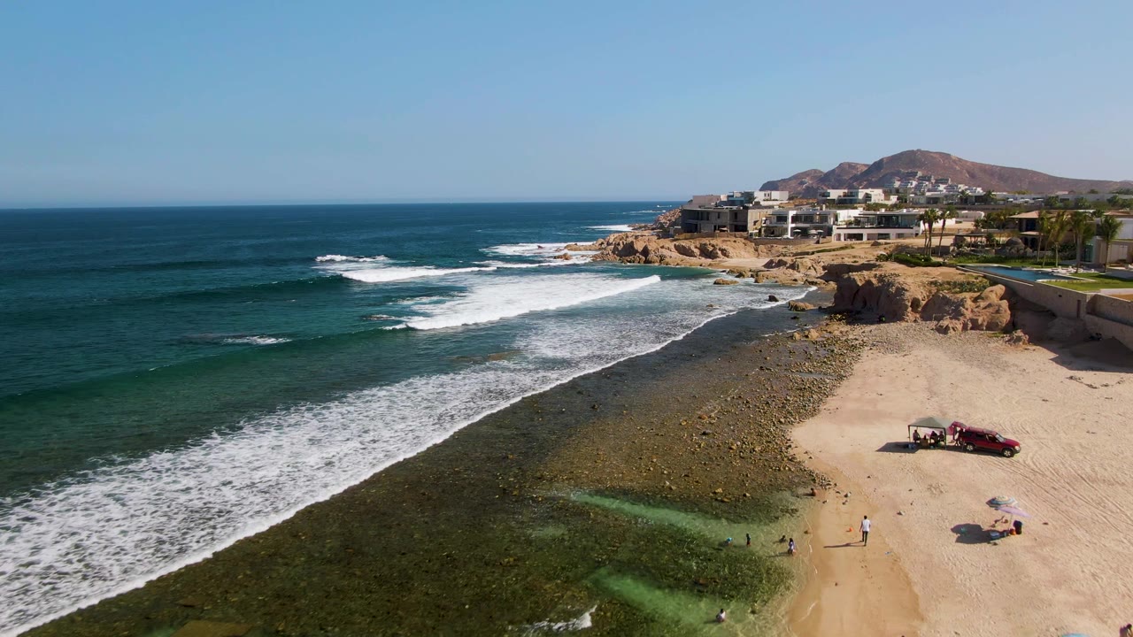 Aerial frame at El Tule Beach in Los Cabos with more ocean filling the left side, foamy surf over the reef, sandy beach on the right, a red vehicle near a small setup, and people spaced along the shoreline.