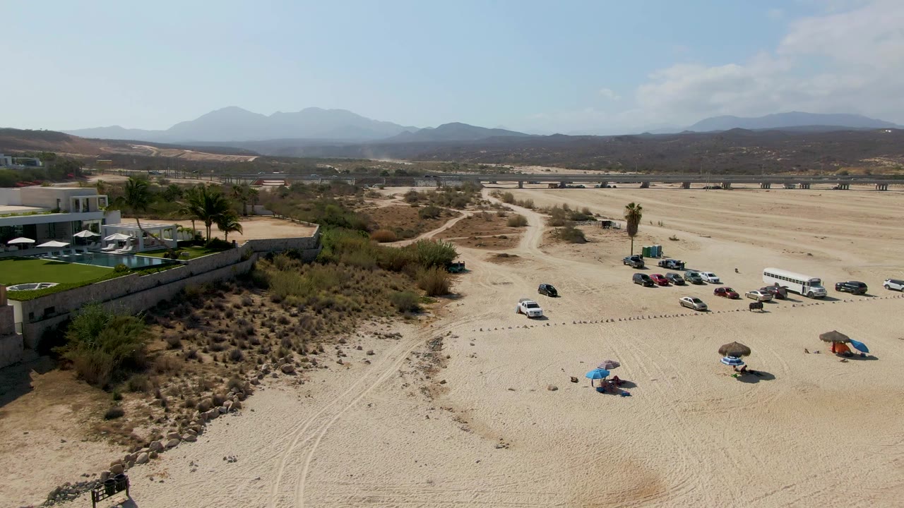 Aerial frame at El Tule Beach in Los Cabos showing a sandy parking area, parked and moving vehicles, umbrellas, a resort building with pool and palms, distant mountains, and no visible people.