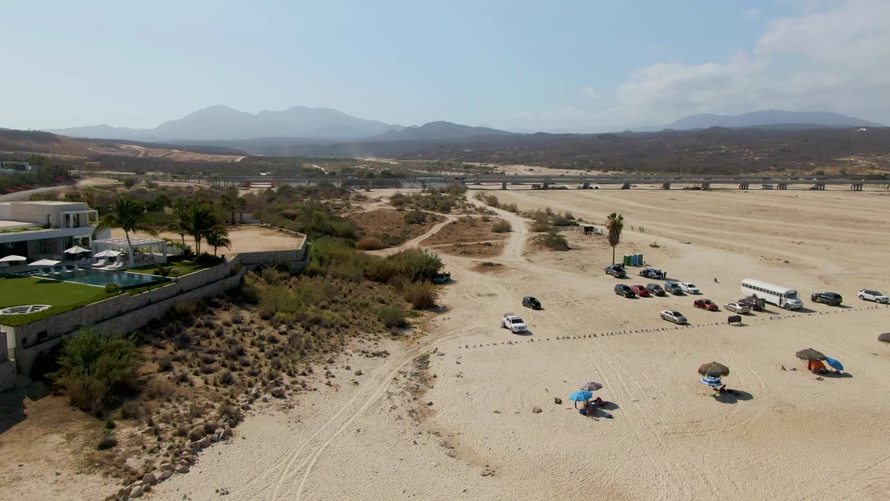 El Tule Beach aerial view with the resort and pool area on the left, vehicle rows across the sand, beach umbrellas in the lower frame, dry scrub, a road, distant mountains, and no visible people.