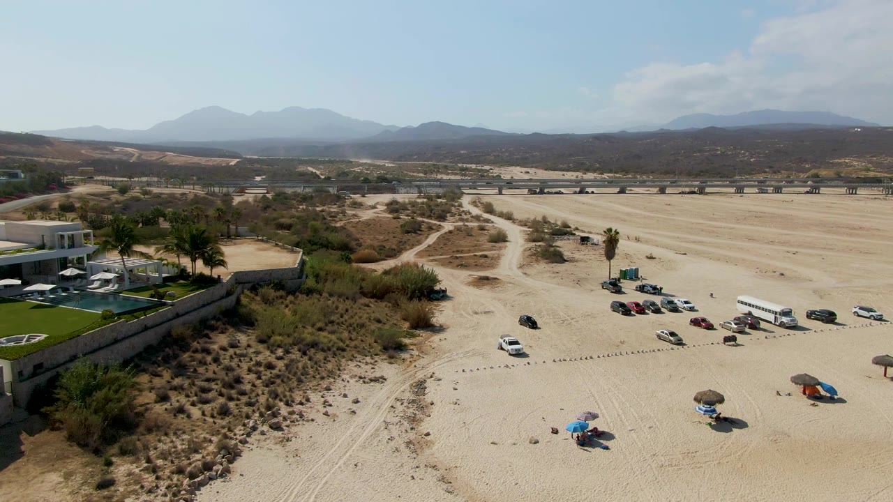 Drone view over El Tule Beach showing vehicles spread across a sandy parking zone, umbrellas near the lower edge, a resort property with palms to the left, highway or road infrastructure in back, mountains, and no visible people.