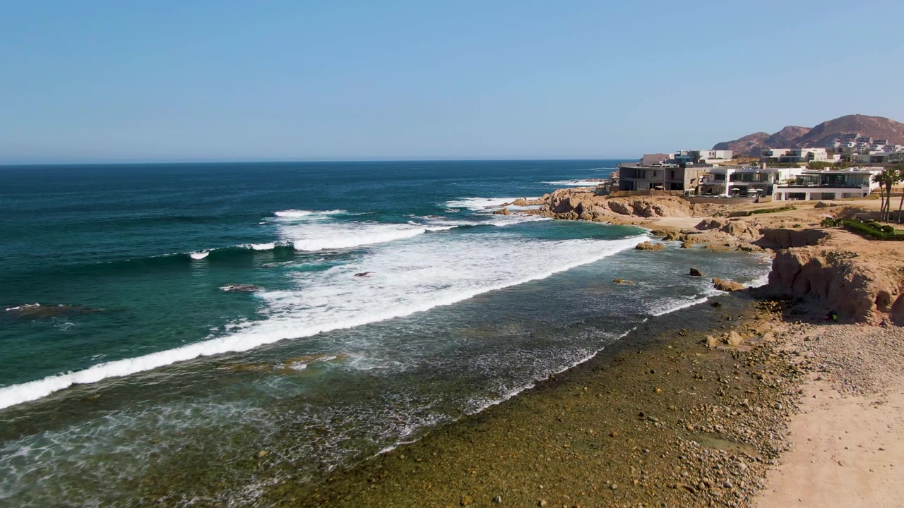 Elevated 4K view of El Tule Beach in Los Cabos with blue ocean, white surf crossing a rocky shelf, sandy cliff edge, distant buildings, and no visible people.
