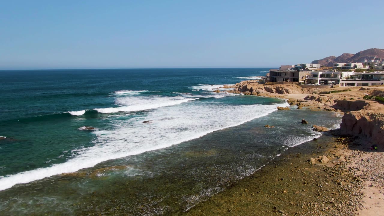 At 0:01, El Tule Beach surf spreads farther across the rocky nearshore water while the horizon, sandy cliff, and cliffside buildings remain visible with no visible people.