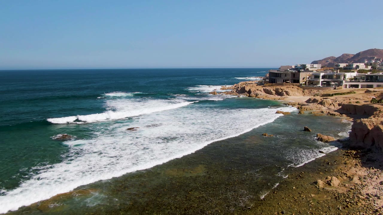 At 0:02, a broad band of whitewater crosses the rocky El Tule Beach shoreline, with deeper blue ocean to the left, cliffside buildings to the right, and no visible people.