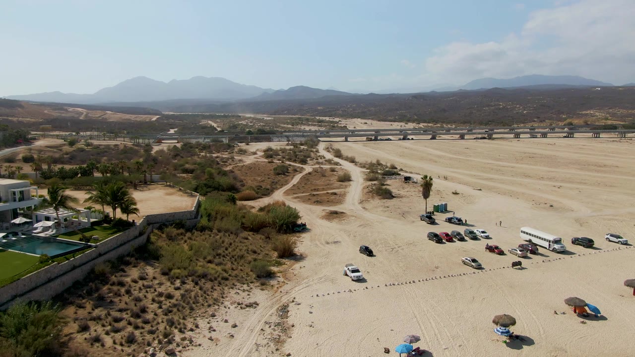 Drone view over El Tule Beach Los Cabos showing sandy open ground, parked cars, a walled pool area with palms, a distant road, mountains, and no visible people.