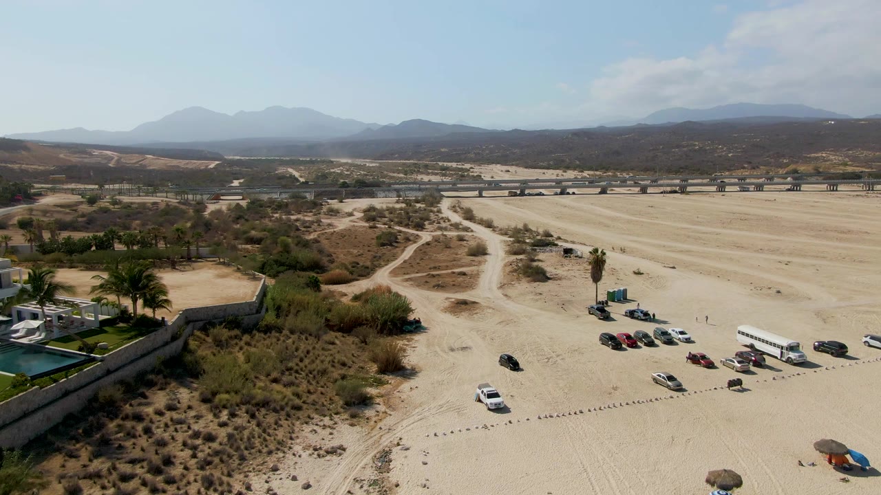 High aerial view of sandy tracks, parked cars, white bus, palms, road, and mountains.