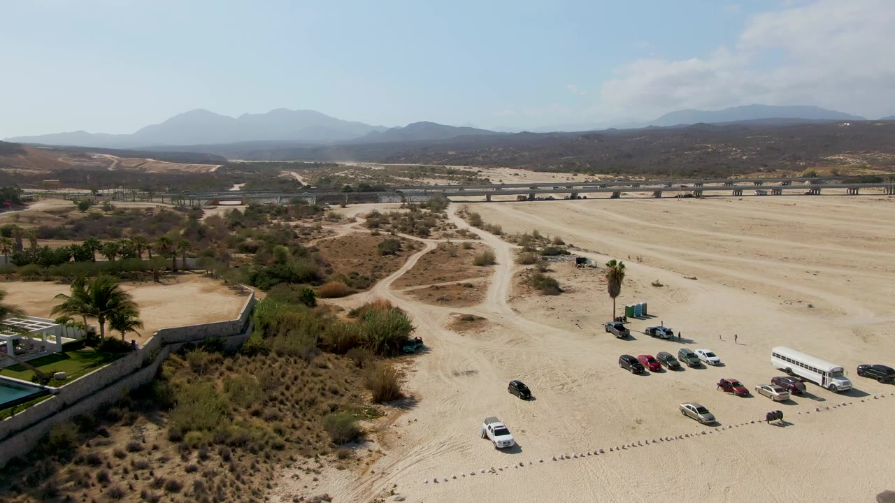 High drone view over El Tule Beach Los Cabos with the parking area shifted toward the right, umbrellas at the lower edge, scrub-covered sand, a long background road, mountains, and no visible people.