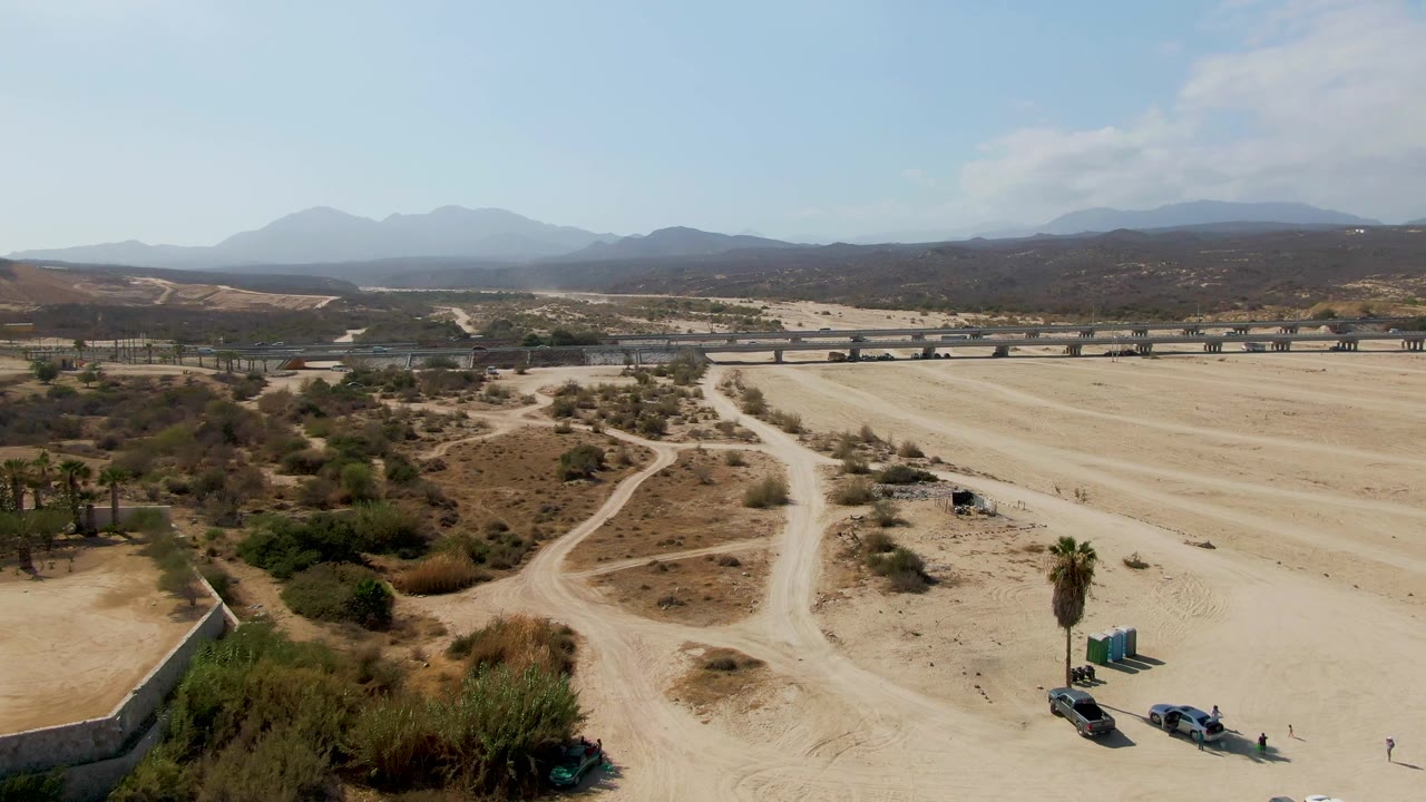 Aerial frame from El Tule Beach Los Cabos showing sandy terrain, winding dirt roads, parked vehicles near the lower edge, a bridge in the midground, distant mountains, and no visible people.