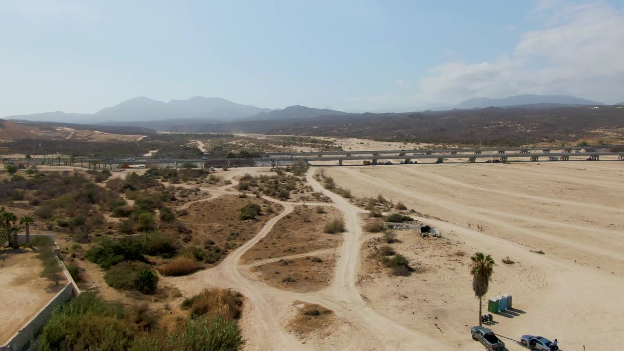 Second aerial frame from El Tule Beach Los Cabos with curving dirt tracks through scrub, vehicles near the lower right, a bridge across the midground, hazy mountains, and no visible people.