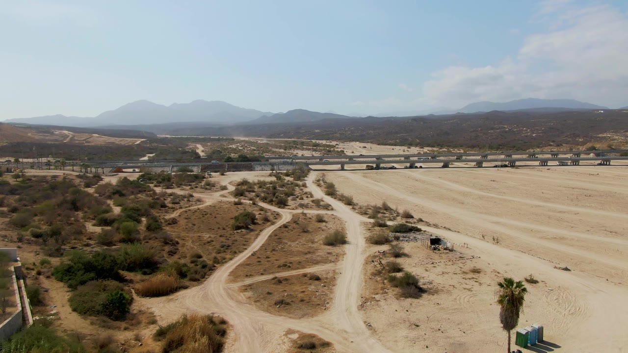 Wide aerial view of sandy open area, dirt road loops, bridge, scrub, and mountains.