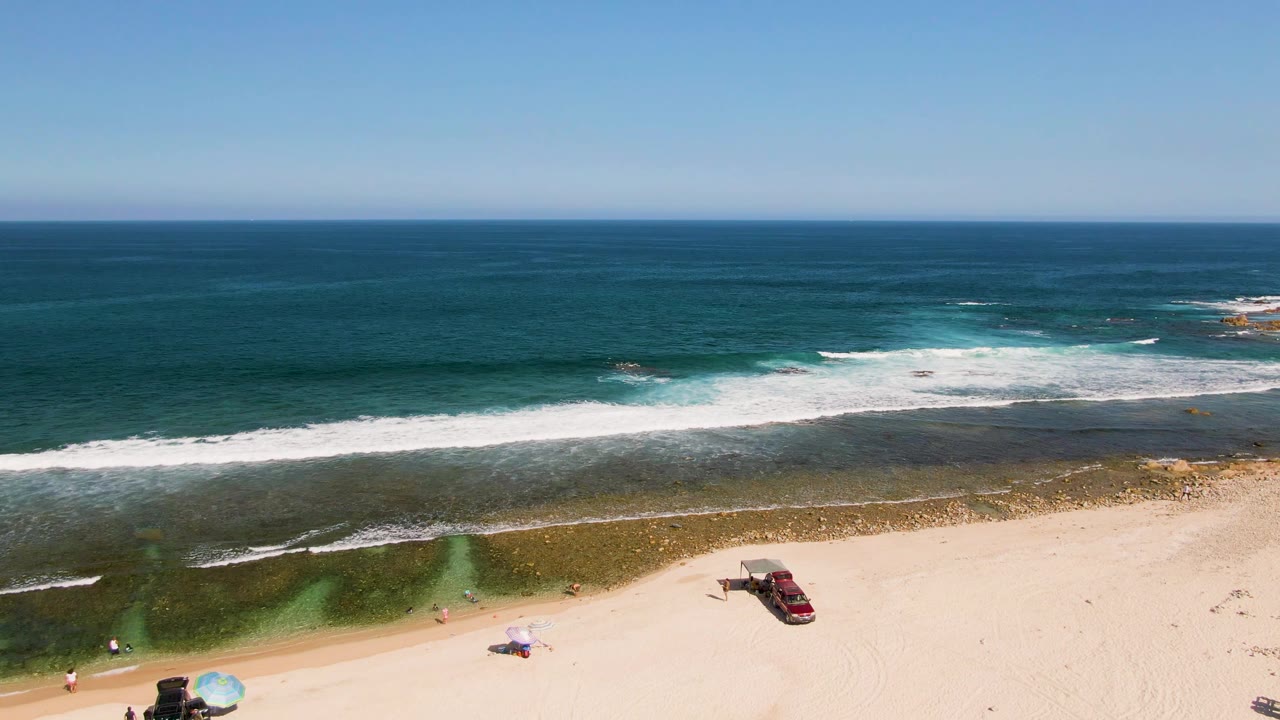 Aerial frame over El Tule Beach in Los Cabos showing blue ocean, white surf, pale sand, a parked red car, a blue umbrella, small beachgoers, and a dark vehicle near the lower edge.