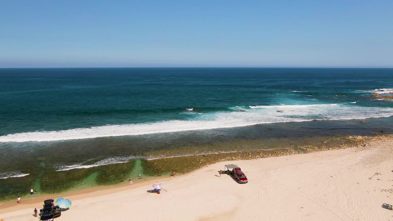 Drone view of surf rolling toward sand with a red car and small beach setup below.