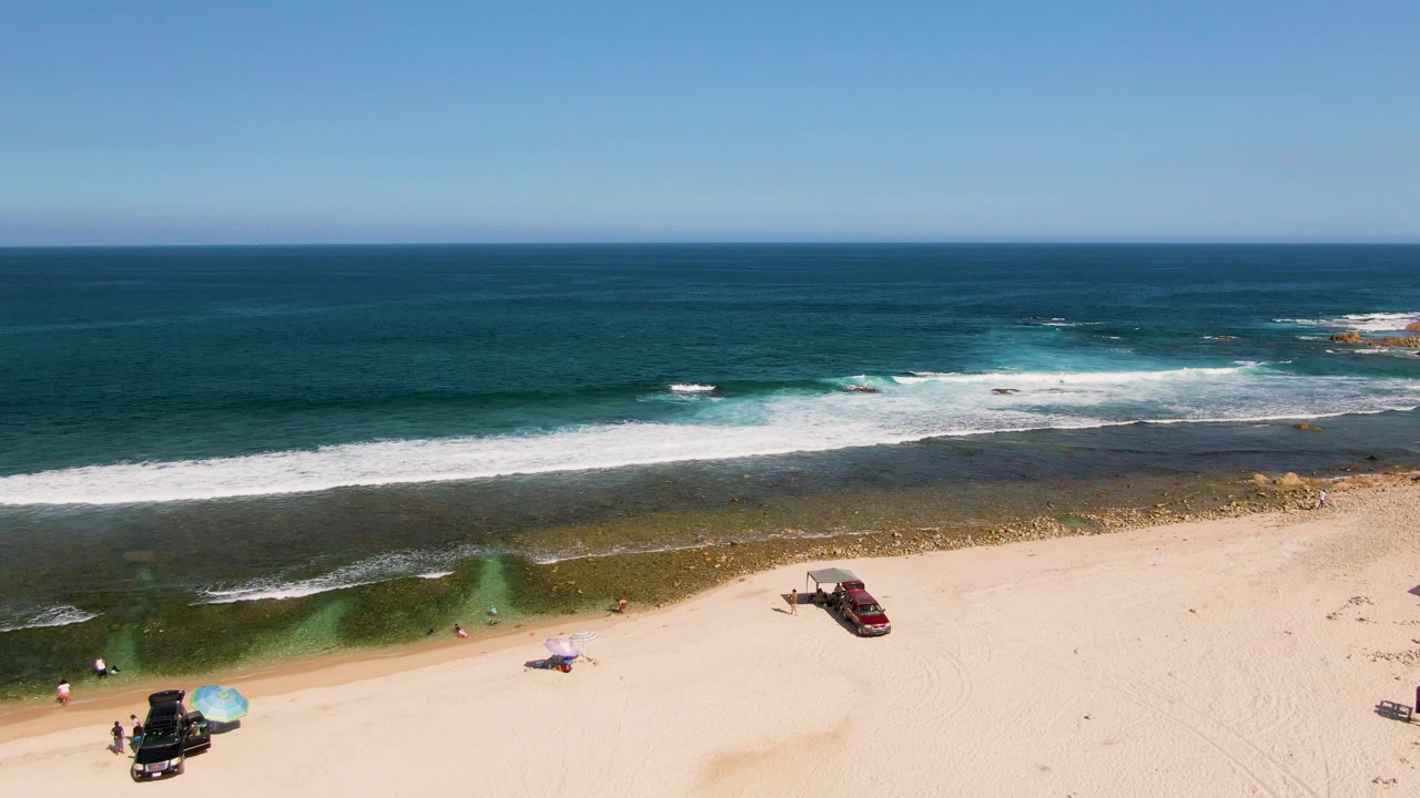 A wide drone frame over El Tule Beach shows the ocean horizon high in frame, bright surf over shallow water, open sand, a red car to the right, and a blue umbrella beside a dark vehicle at the bottom.