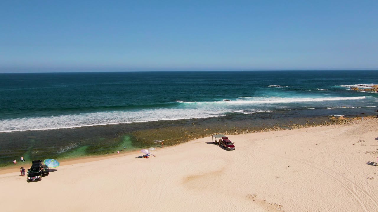 Aerial view of El Tule Beach in Los Cabos with two parked vehicles, small beachgoers, blue umbrellas near the sand, white surf, and a clear ocean horizon.