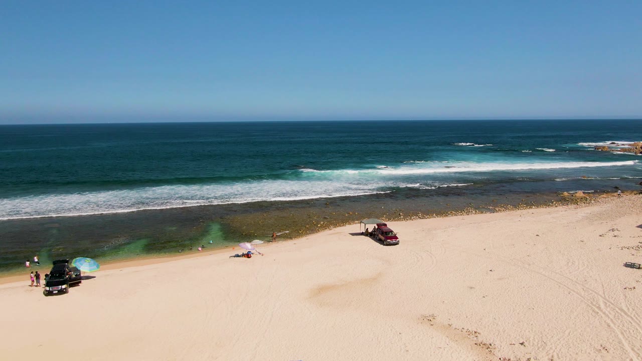 Aerial shoreline view with beachgoers near umbrellas and vehicles beside rolling surf.