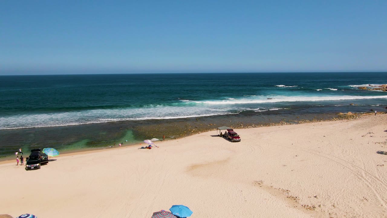 Aerial frame at El Tule Beach with a broad sweep of sand, visible people near umbrellas, a dark vehicle close to the shoreline, a red vehicle farther right, and layered surf beyond.
