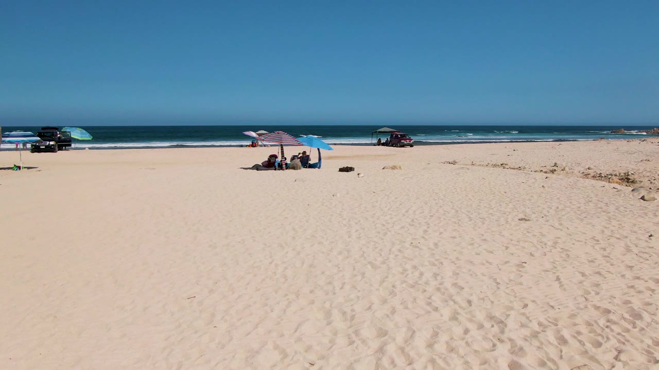 Wide view of El Tule Beach in Los Cabos with open sand, seated beachgoers under umbrellas, parked vehicles near the shoreline, low ocean waves, and a clear blue sky.