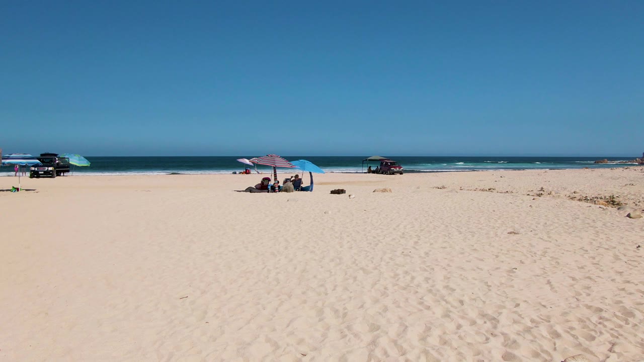Beachgoers sit under umbrellas on a wide sand beach with vehicles near the surf.