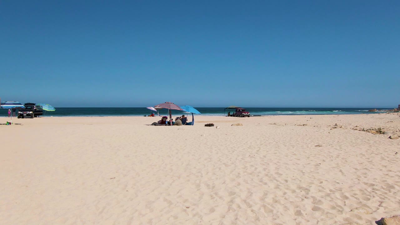 El Tule Beach view with beachgoers under umbrellas slightly left of center, a dark vehicle near the water, a red vehicle farther right, and rocks along the sandy dune edge.