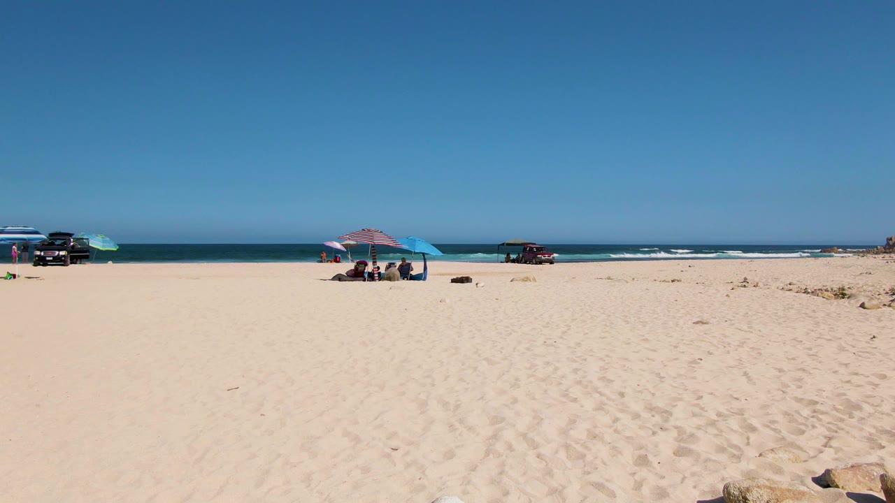Opening view of El Tule Beach in Los Cabos with broad sand in the foreground, small beach umbrellas and seated people near the waterline, parked vehicles, and low waves under clear sky.