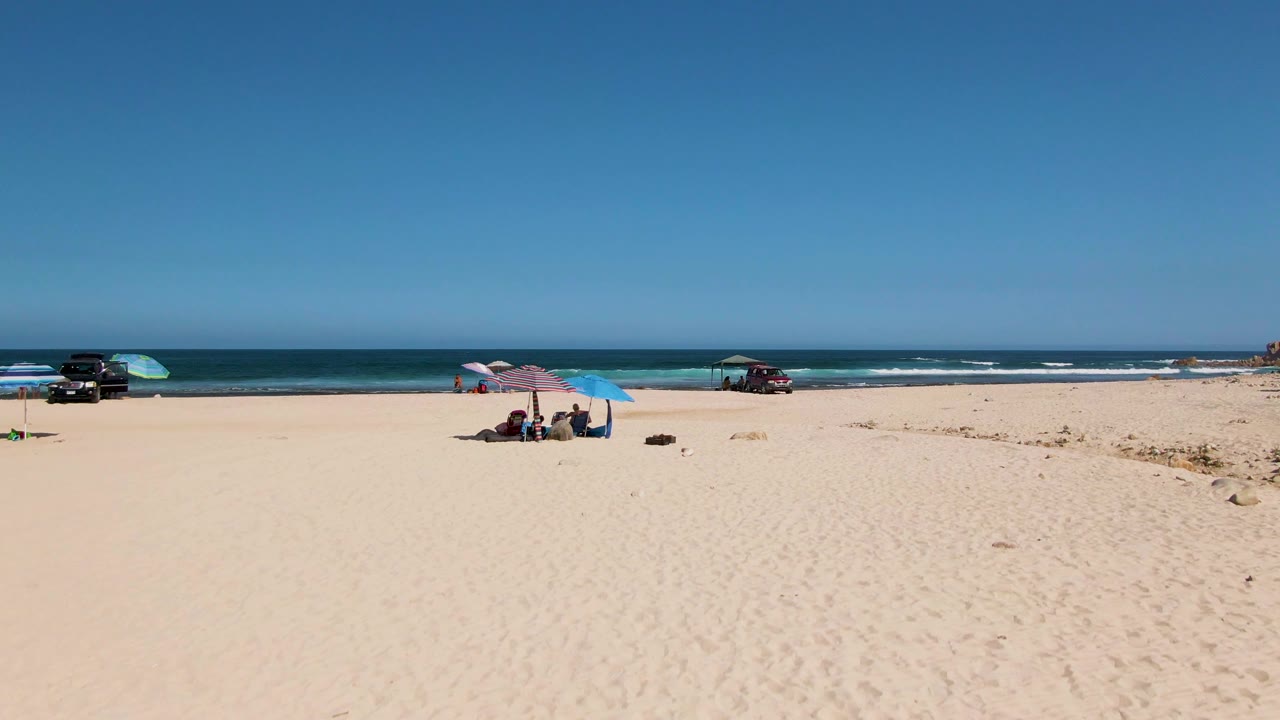 Sunny beach scene with two umbrellas, seated people, vehicles, waves, and clear horizon.