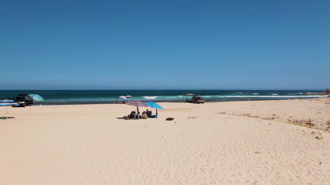 El Tule Beach view with a larger stretch of empty sand in front, beachgoers sitting beneath pink and blue umbrellas, vehicles near the shoreline, and white surf lines in the background.