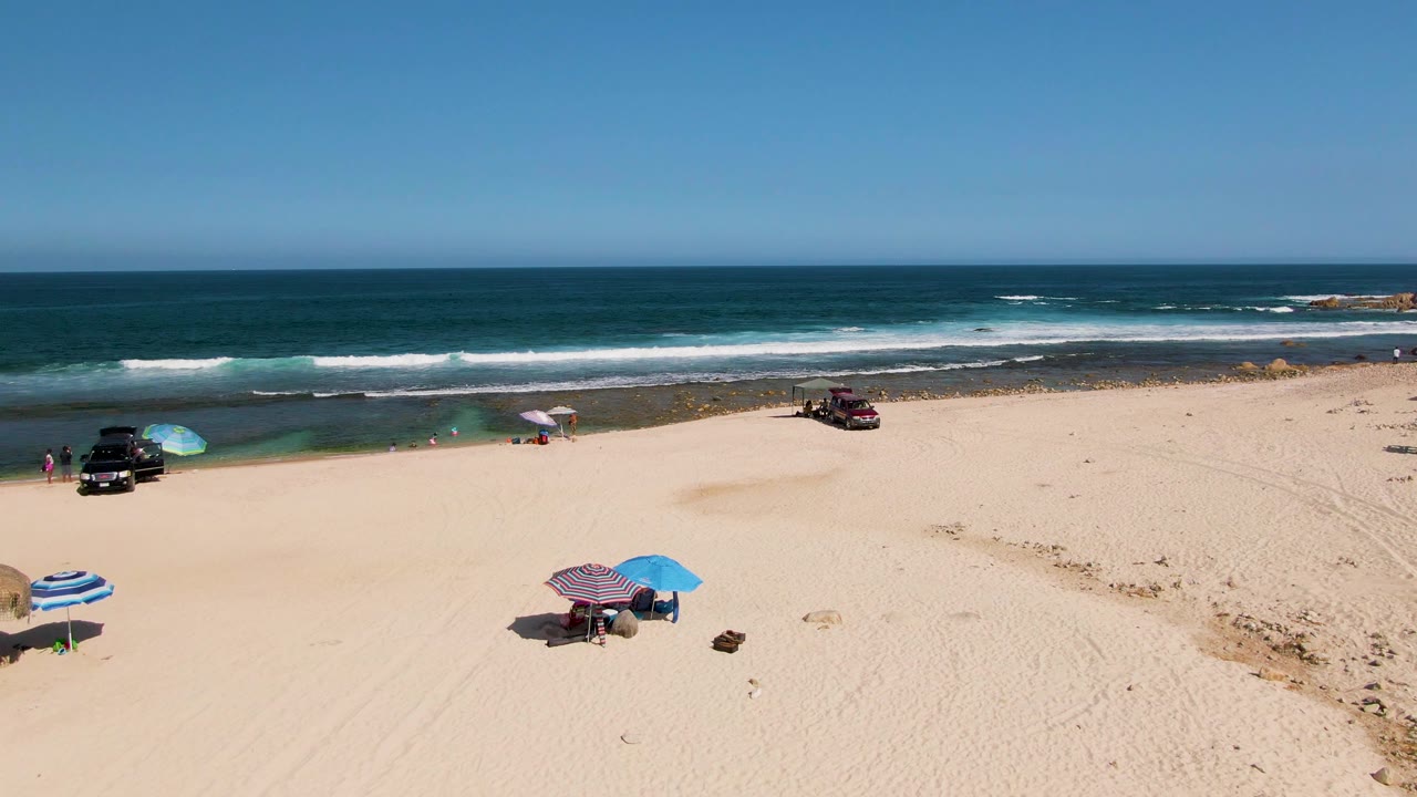 Wide aerial frame at El Tule Beach in Los Cabos, Mexico, showing open sand, small umbrella setups, parked vehicles near the waterline, white surf, and a clear horizon.