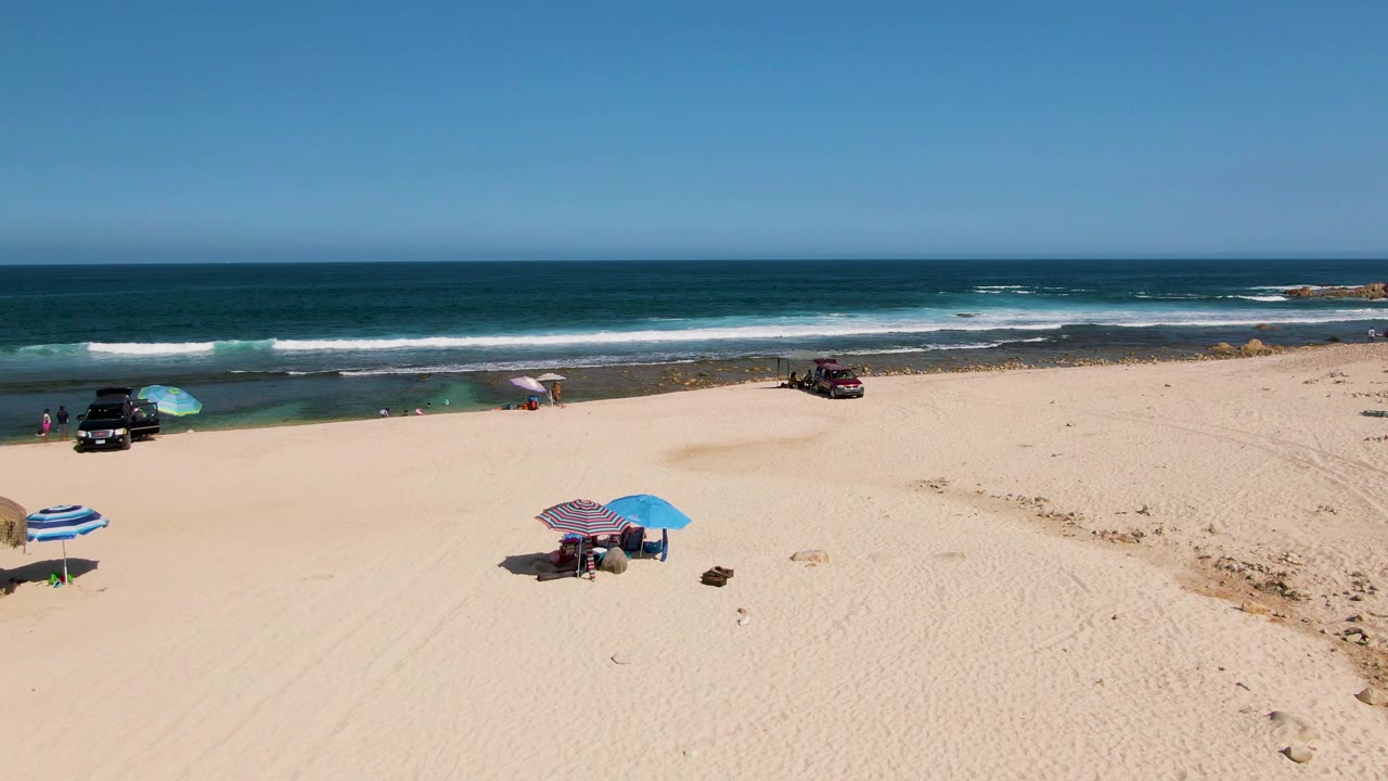 Aerial beach frame with striped and blue umbrellas, people on sand, vehicles, and waves.
