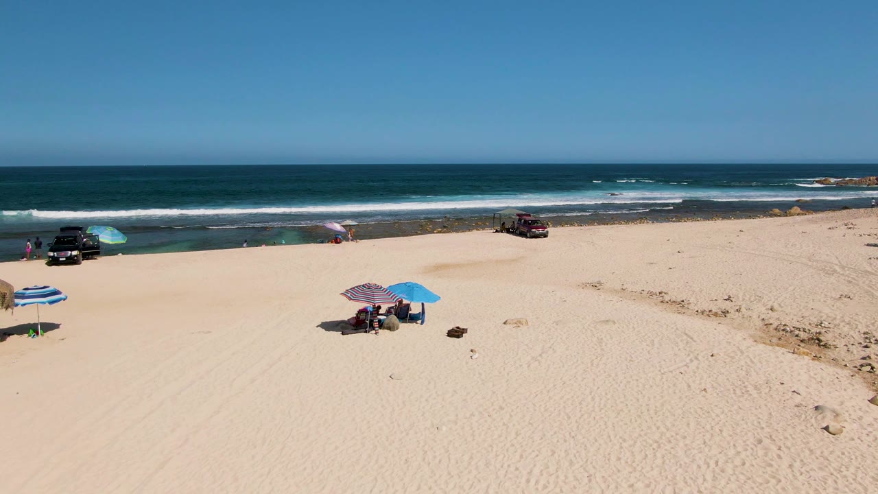 High wide view at El Tule Beach in Los Cabos, Mexico, showing two umbrella areas separated by open sand, dark vehicles near the shoreline, and surf bands behind them.