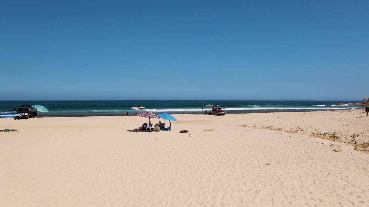 Wide view of El Tule Beach in Los Cabos with pale sand in front, beachgoers under blue and striped umbrellas, parked vehicles near the surf, and a flat blue horizon.