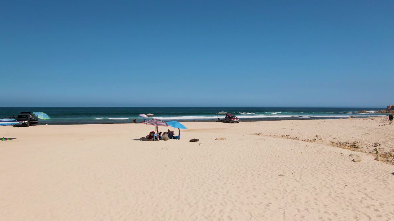 Beachgoers sit under umbrellas on a sandy shore with ocean waves behind them.