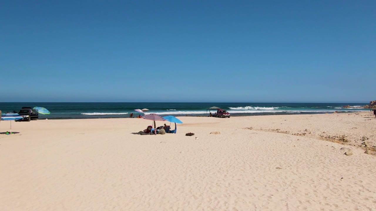 The El Tule Beach view holds a broad sandy foreground, seated beachgoers under two umbrellas, vehicles close to the water, and white surf moving along the blue ocean.