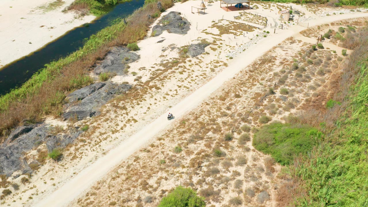 Aerial frame in San Jose Del Cabo with the small vehicle farther along the sandy road, the dark waterway at left, dry tan vegetation at center, and no visible people.