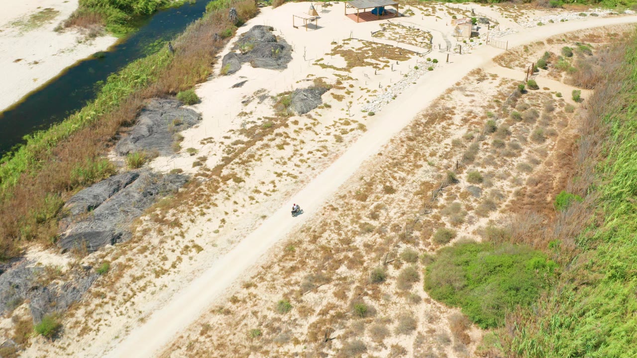Aerial frame in San Jose Del Cabo showing the vehicle near the center-right stretch of the sandy road, with rocks, scrub, green reeds, and a narrow waterway nearby and no visible people.