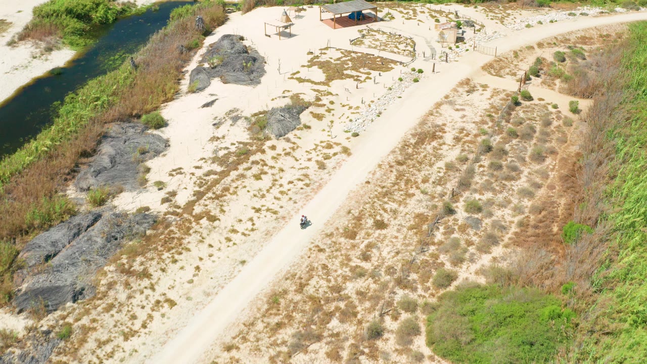 Aerial frame in San Jose Del Cabo with the vehicle lower on the diagonal sandy road, dark water at left, rocky ground and sparse scrub in the middle, and no visible people.