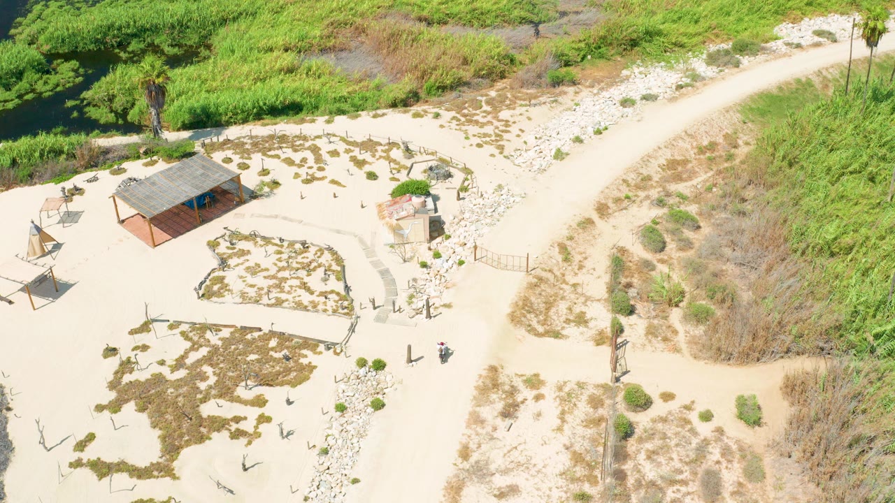 Aerial frame from San Jose Del Cabo showing a pale sandy path curving past a small roofed building, scattered scrub, posts, and green vegetation at the edge of the frame.