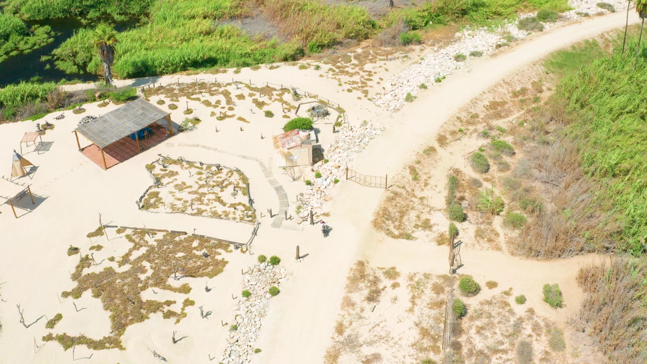 Aerial frame from San Jose Del Cabo showing the curving dirt road more prominently on the right, with the small roofed structure, scattered plants, and pale sand filling the left side.