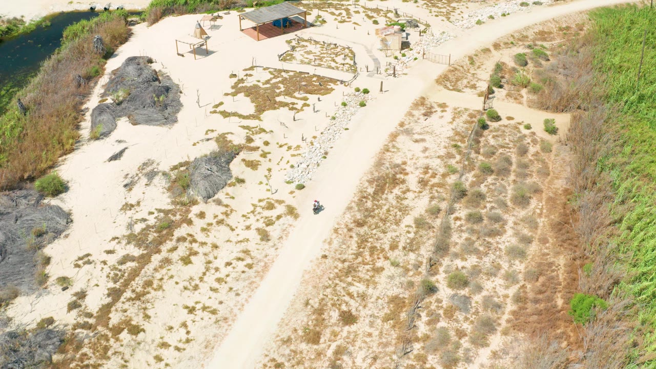 High aerial frame over a sandy dirt road, dark rock outcrops, scrub vegetation, and small shade structures in San Jose Del Cabo, with no visible people.