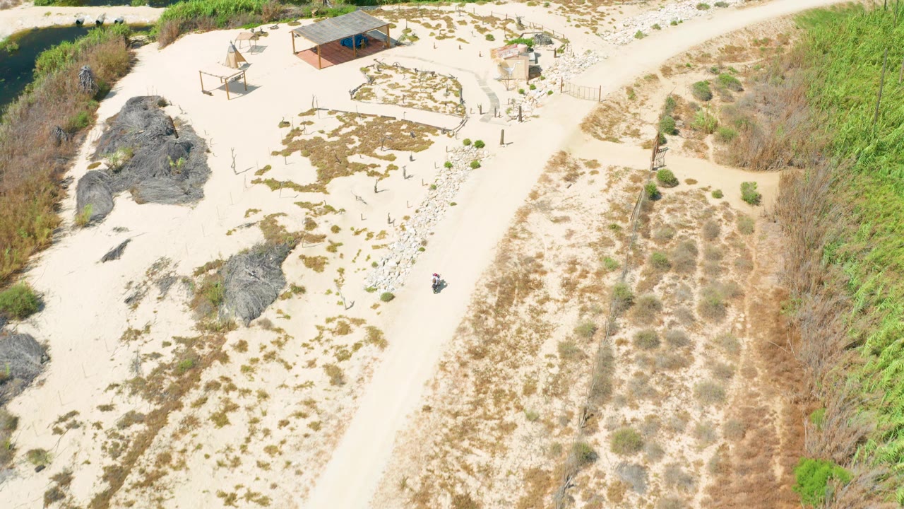 Aerial frame in San Jose Del Cabo showing the pale dirt road curving past sandy lots, dark rocks, green brush, and a small roofed structure, with no visible people.