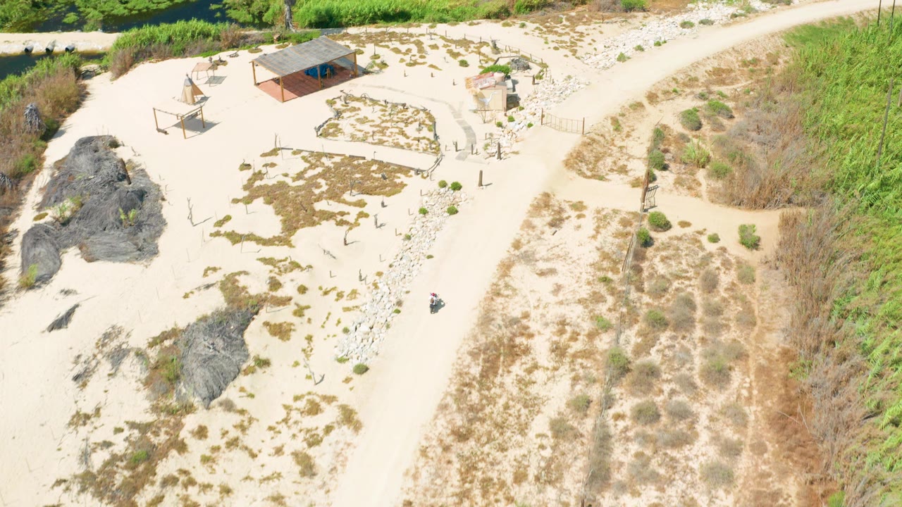 High overhead frame of the San Jose Del Cabo coastal road area with sandy terrain, a roofed structure, shade frames, dark rocks, dry brush, and no visible people.