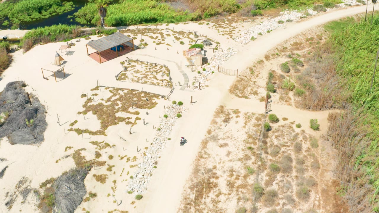 Aerial view of a small roofed structure beside sand, rocks, and a dirt road.