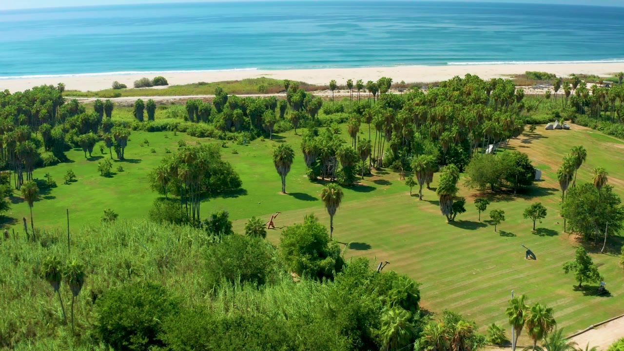 Elevated San Jose Del Cabo frame with bright lawn, clustered trees, small park features, a narrow sand strip, and blue ocean bands beyond.