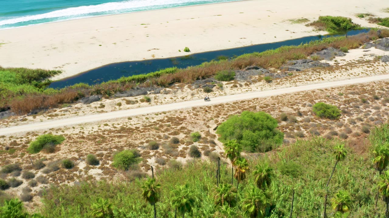 Aerial view of a beach, water channel, coastal road, scrub, palms, and a small vehicle.