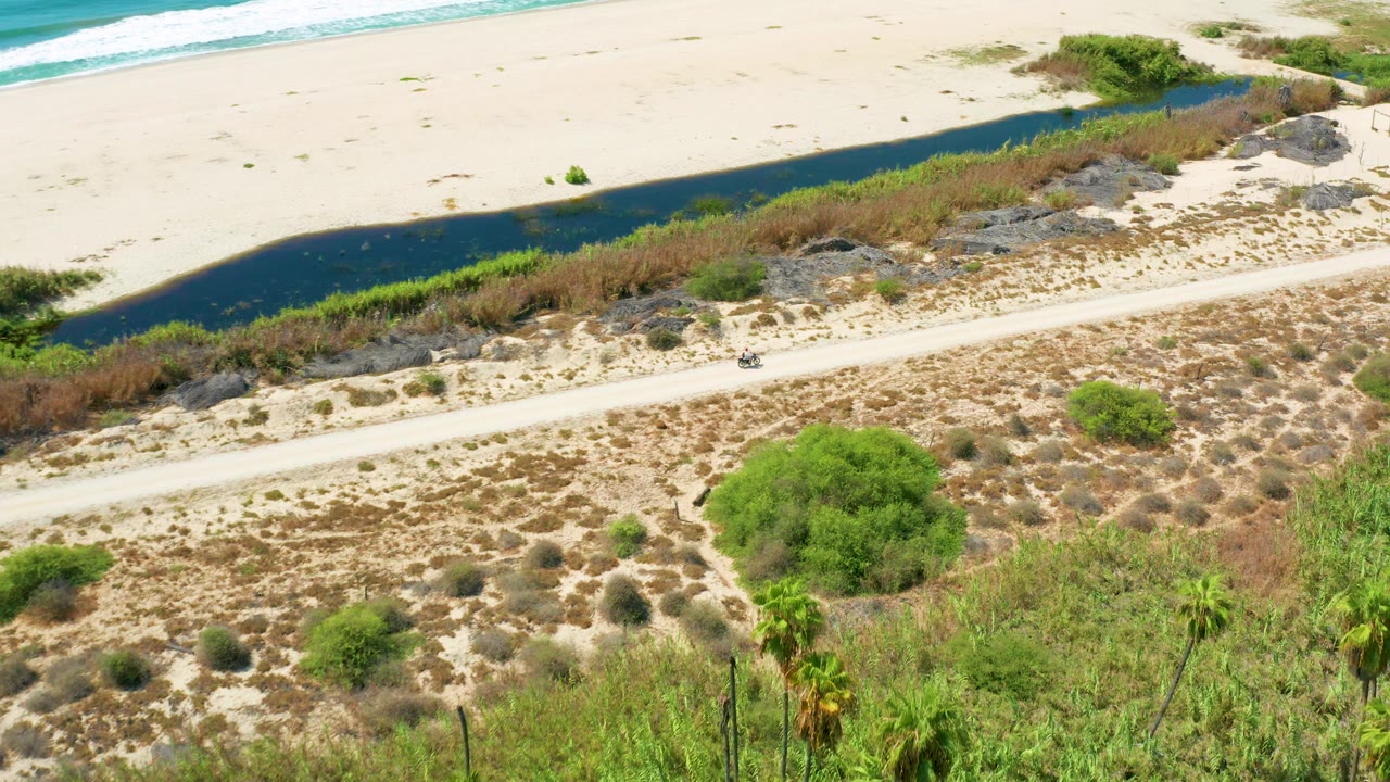 Aerial view in San Jose Del Cabo with the road crossing the middle of frame, a dark channel above it, ocean surf beyond the beach, palms at the bottom edge, and no visible people.