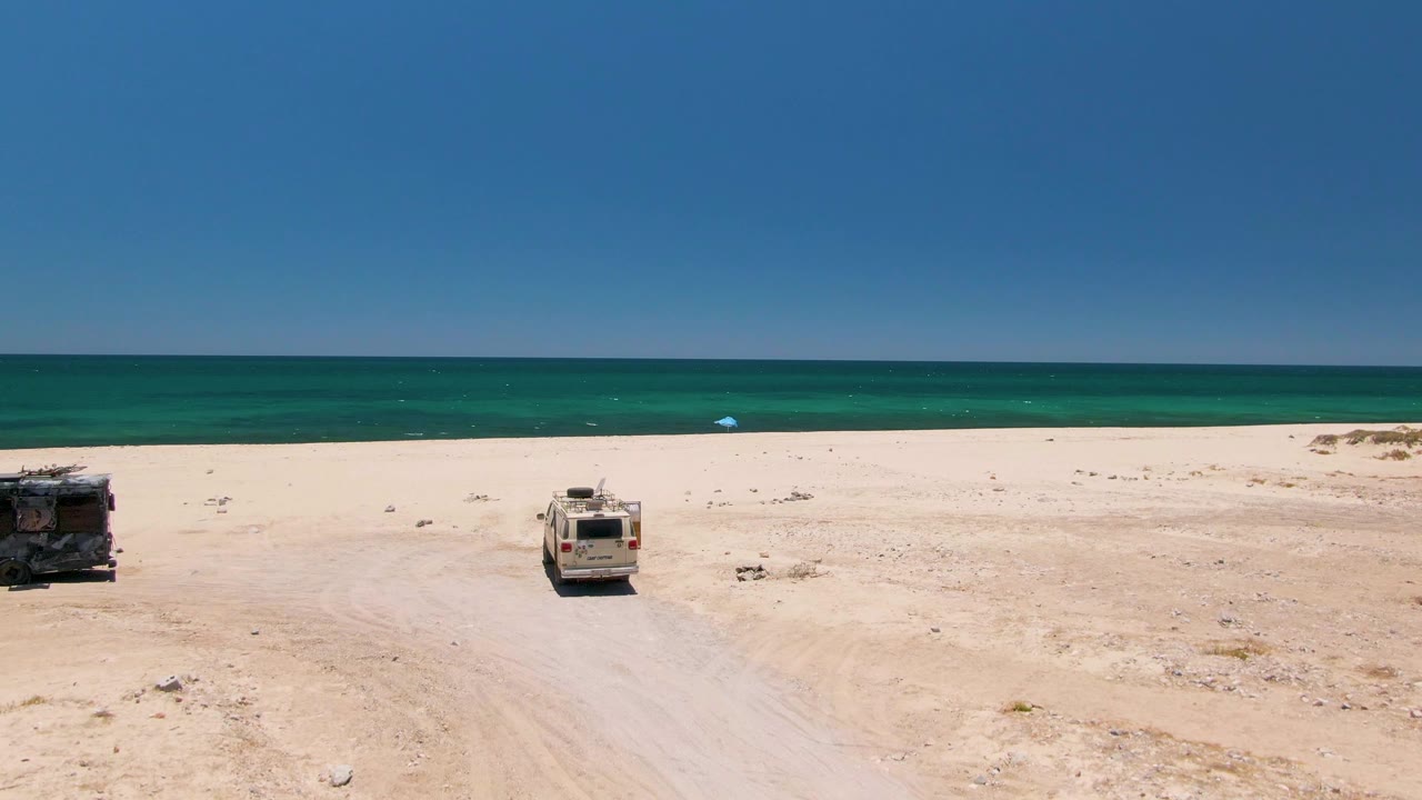 Aerial view at Playa Miramar Los Cabos showing two parked vehicles on pale sand beside turquoise water and clear sky, with no visible people.
