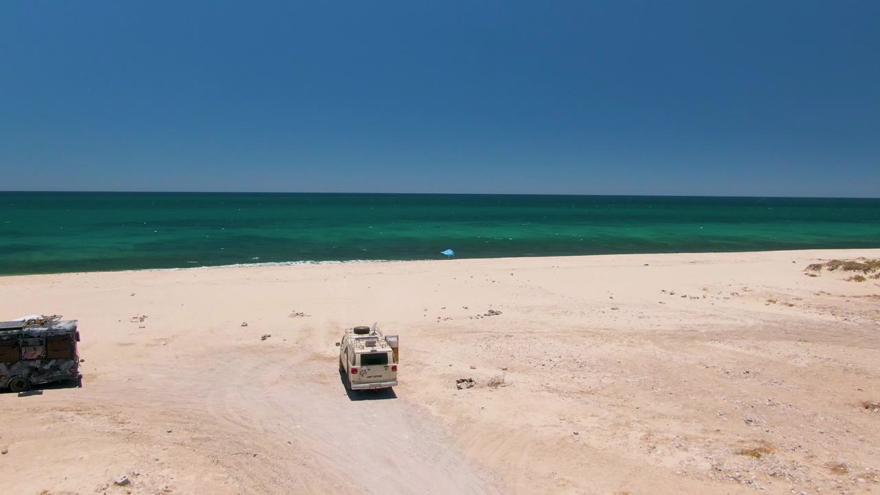Aerial frame over Playa Miramar Los Cabos with the vehicles slightly farther into view on the sand and the turquoise ocean stretching across the upper frame, with no visible people.